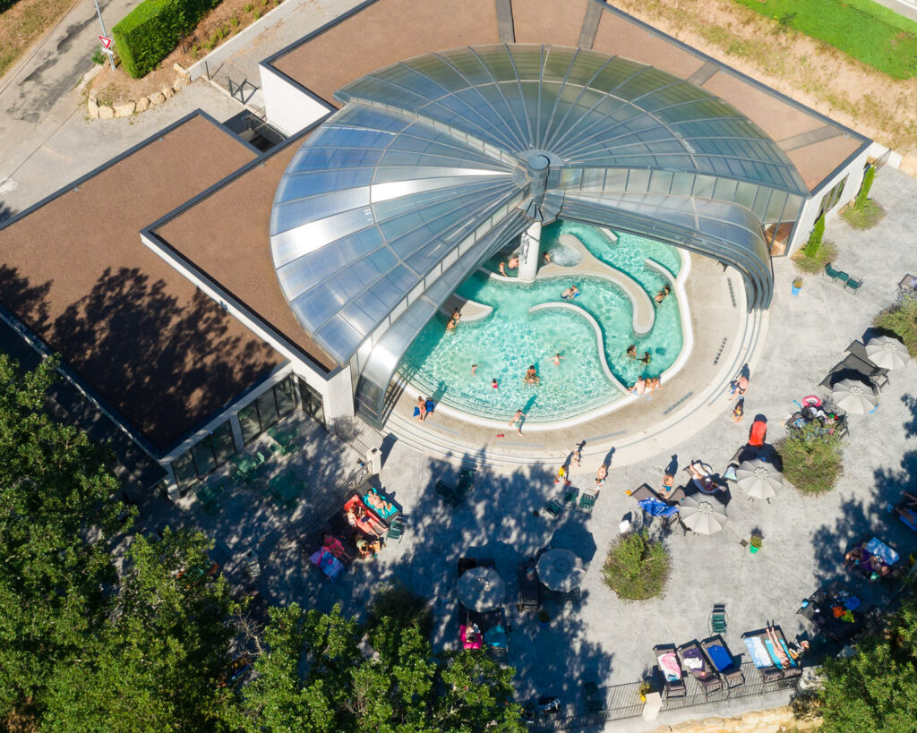 Vue aérienne d'une piscine extérieure couverte d'un dôme transparent avec toboggan aquatique, baigneurs dans l'eau et visiteurs sur la terrasse environnante.