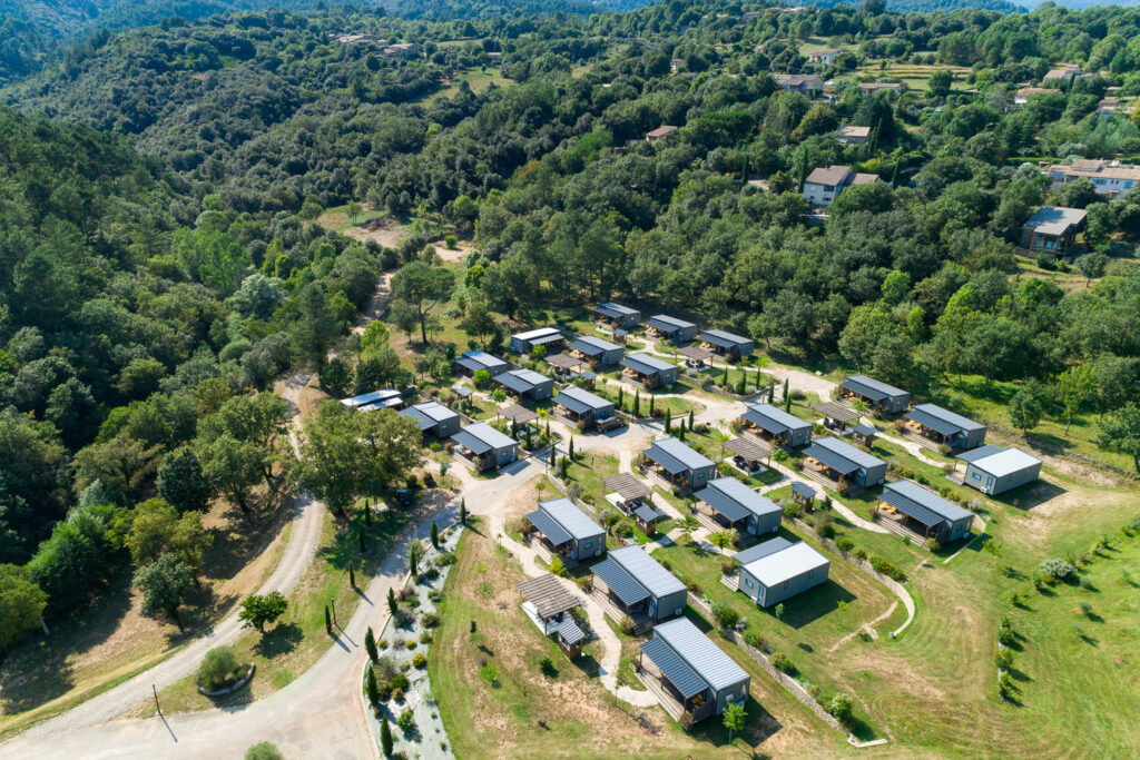 Vue aérienne d'un village de chalets modernes avec toits gris disposés en rangées, entouré de forêts verdoyantes et de collines.