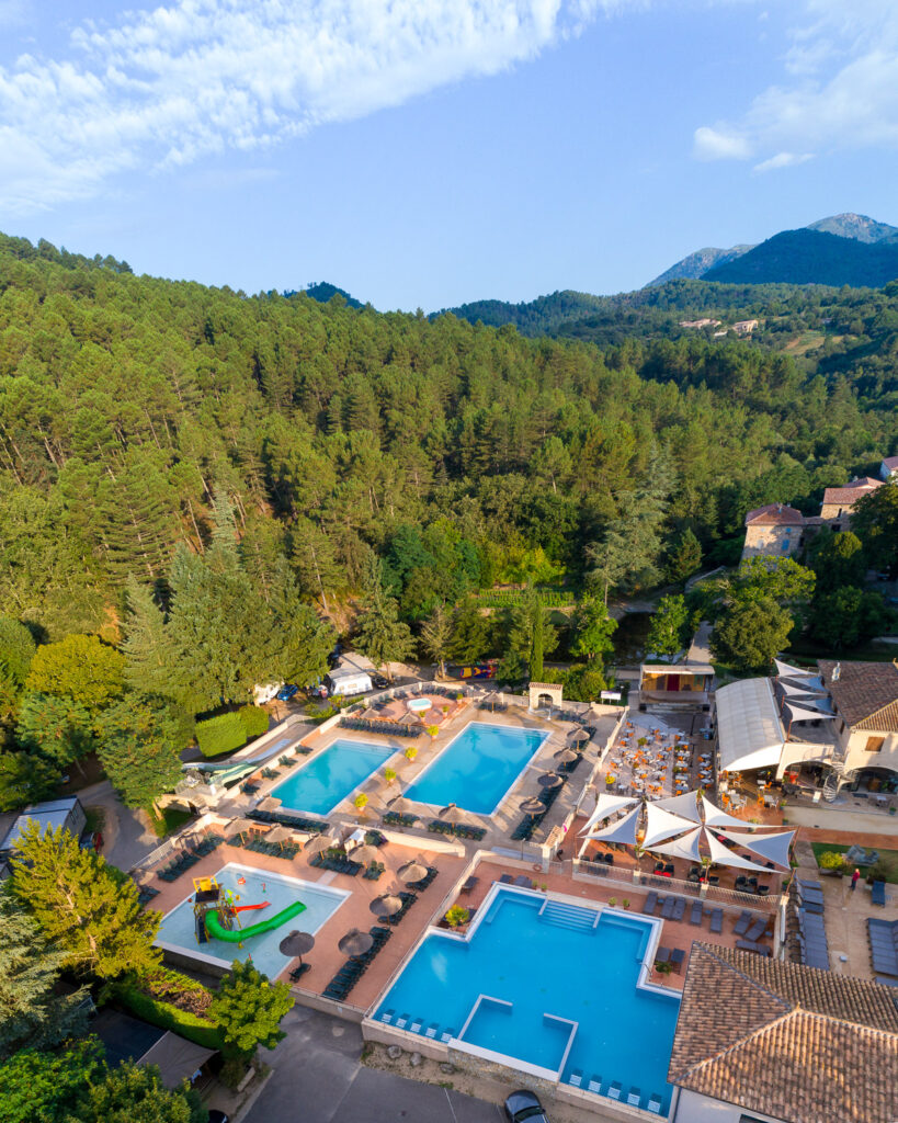 Vue aérienne d'un complexe de camping en Ardèche avec trois piscines, pataugeoire et restaurant, niché dans une vallée boisée aux montagnes en arrière-plan.