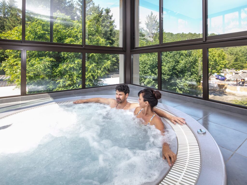 Couple se détendant dans un jacuzzi intérieur avec grande baie vitrée offrant vue sur jardin verdoyant et ciel bleu.