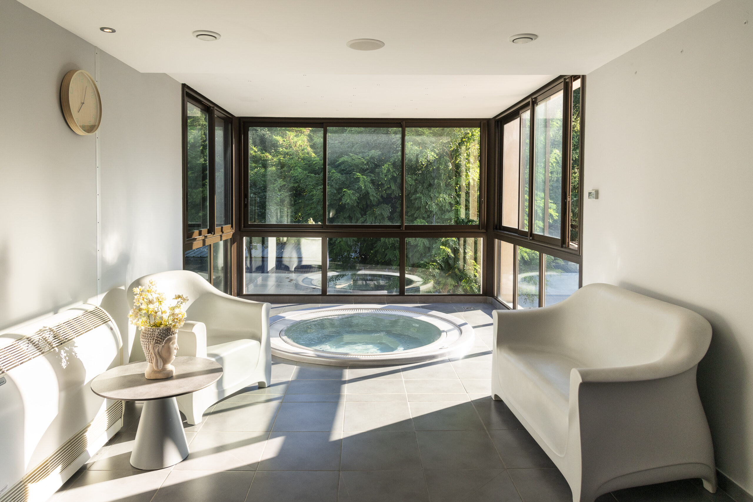 Espace spa intérieur lumineux avec deux fauteuils blancs, une petite table, une vasque à bulles, et une grande baie vitrée donnant sur la verdure.