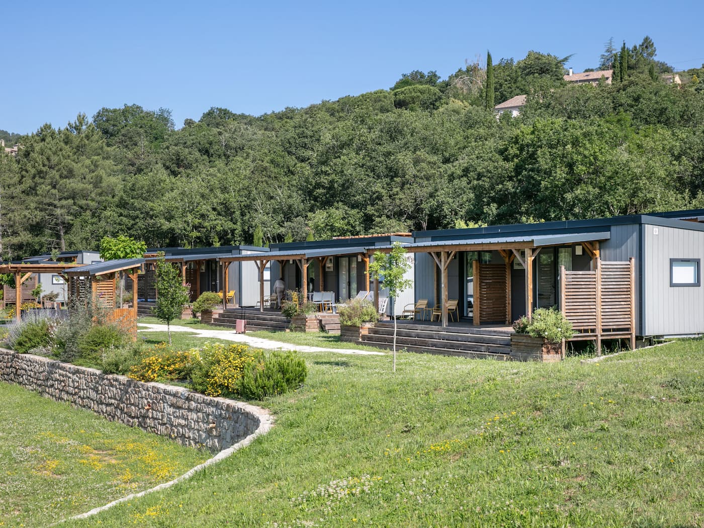 Groupe de maisons modulaires modernes en conteneurs gris et blancs, avec terrasse en bois, fauteuils et escaliers, dans un jardin verdoyant bordé de murets et entouré de collines boisées, sous un ciel bleu.