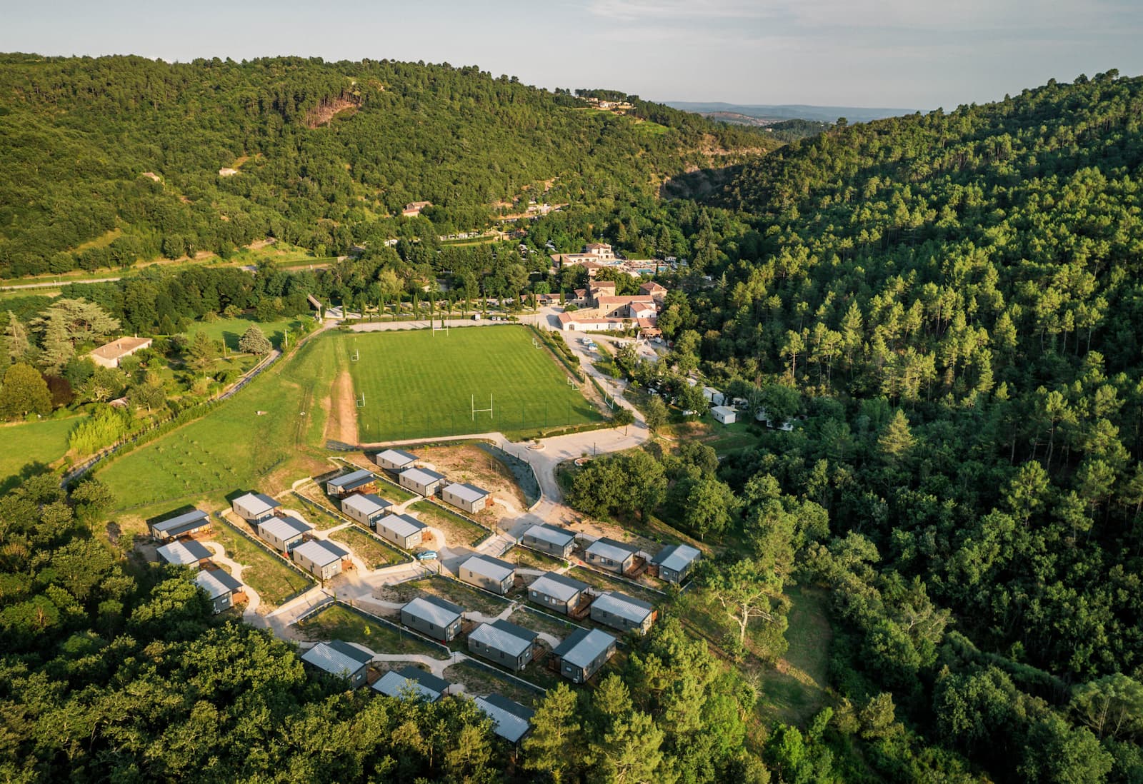 Vue aérienne d’un petit lotissement avec des maisons et dépendances au milieu de champs et de collines boisées, au bord d’une vallée, sous un ciel clair.