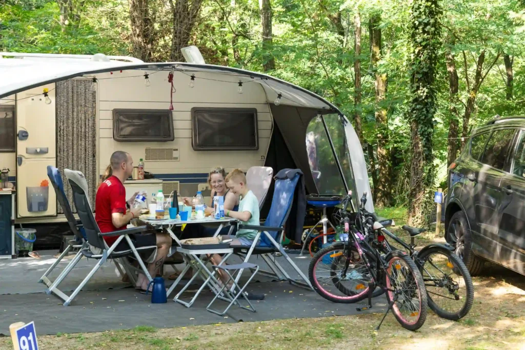 Famille en camping déjeunant dehors devant leur caravane équipée d'un auvent, avec vélos d'enfant au premier plan dans une forêt verdoyante.